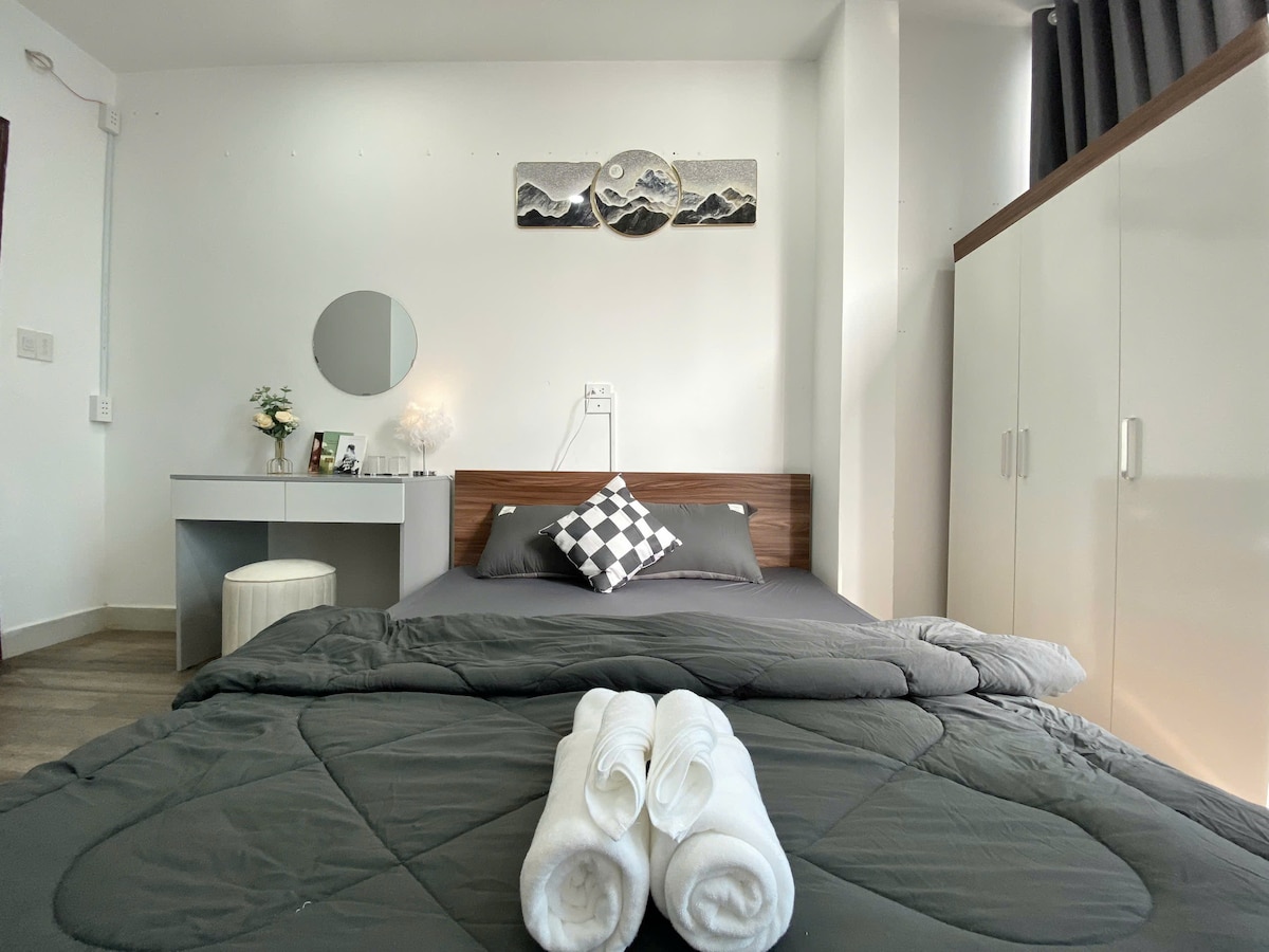 A comfortable sleeping area is presented with a spacious bed dressed in dark linens. A simple, stylish table with a mirror is positioned beside the bed. Neatly rolled towels are placed on the bed, while a wardrobe occupies the corner, enhancing the room's functionality.