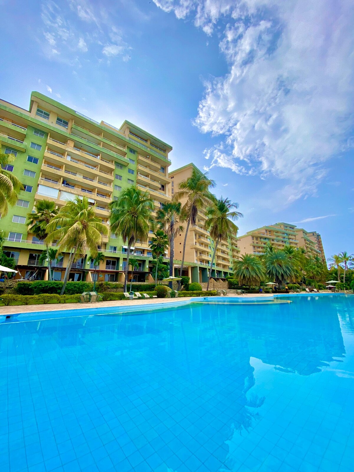 A sparkling blue swimming pool is surrounded by well-maintained gardens and palm trees, with several lounge chairs placed along the water's edge. The multi-story buildings, featuring a mix of green and beige facades, provide a backdrop against the clear sky.