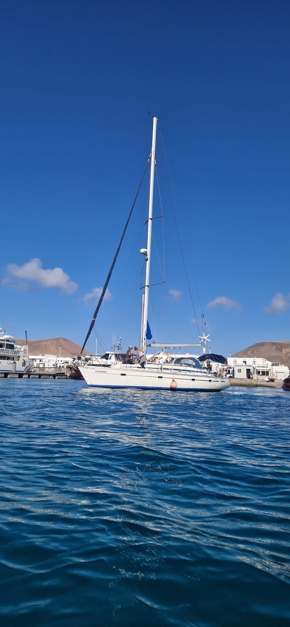 A sailboat is anchored in calm waters, featuring a tall mast and a bright blue sail. The background includes a marina with several boats and distant hills under a clear blue sky, reflecting a serene coastal atmosphere.