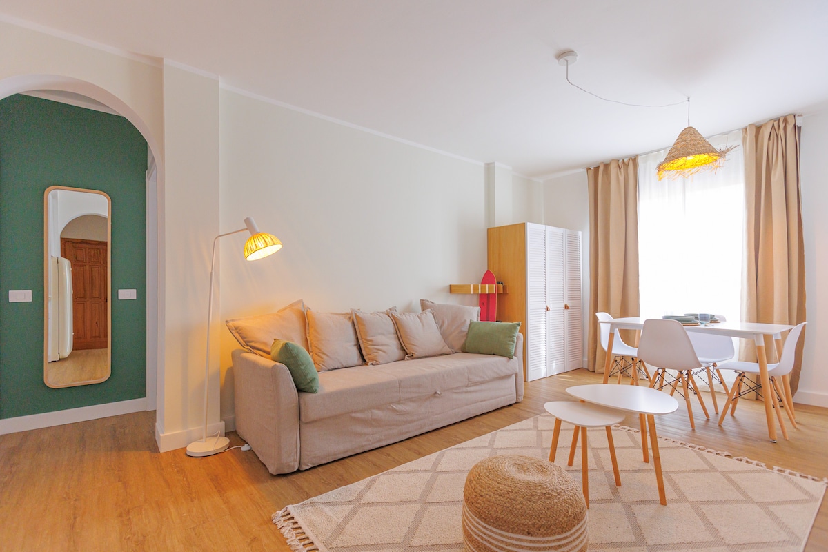 A light-filled living area features a neutral-toned sofa adorned with cushions, complemented by a round coffee table on a textured rug. A dining table with white chairs is positioned nearby, while a decorative pendant light hangs above, adding warmth to the space. A large mirror reflects the room.