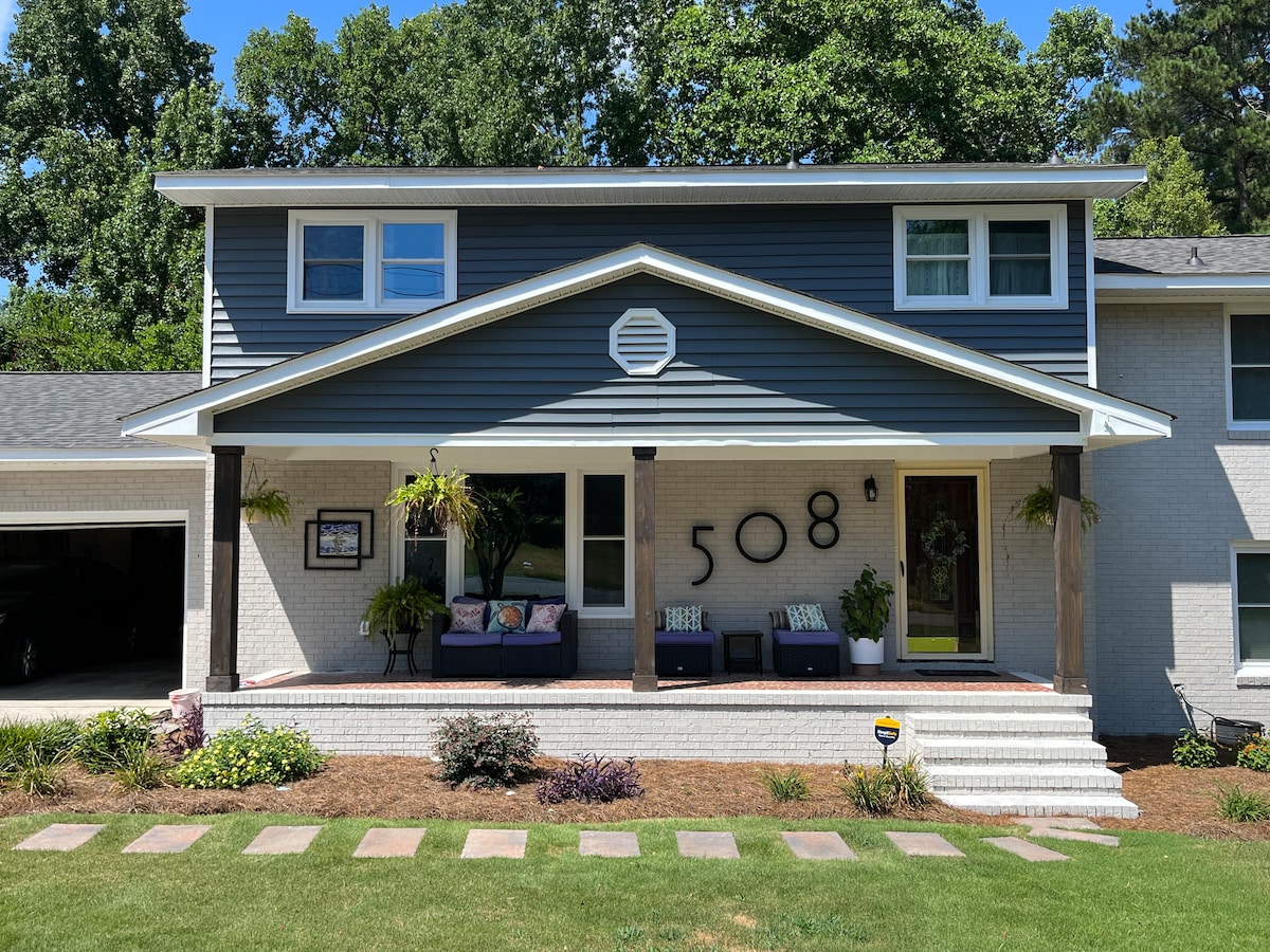 The exterior of the house features a modern two-story design with a mix of blue and white siding. A welcoming porch is adorned with seating and potted plants. Stone steps lead to the entryway, while a well-maintained lawn and pathway complement the landscape.
