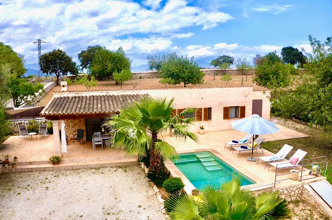 The image captures a rural property surrounded by greenery, featuring a private salt-water pool. The outdoor area includes shaded seating and sun loungers alongside the pool. A smooth pathway leads to the entrance of the house, complemented by a few well-placed palm trees and open views of the landscape.