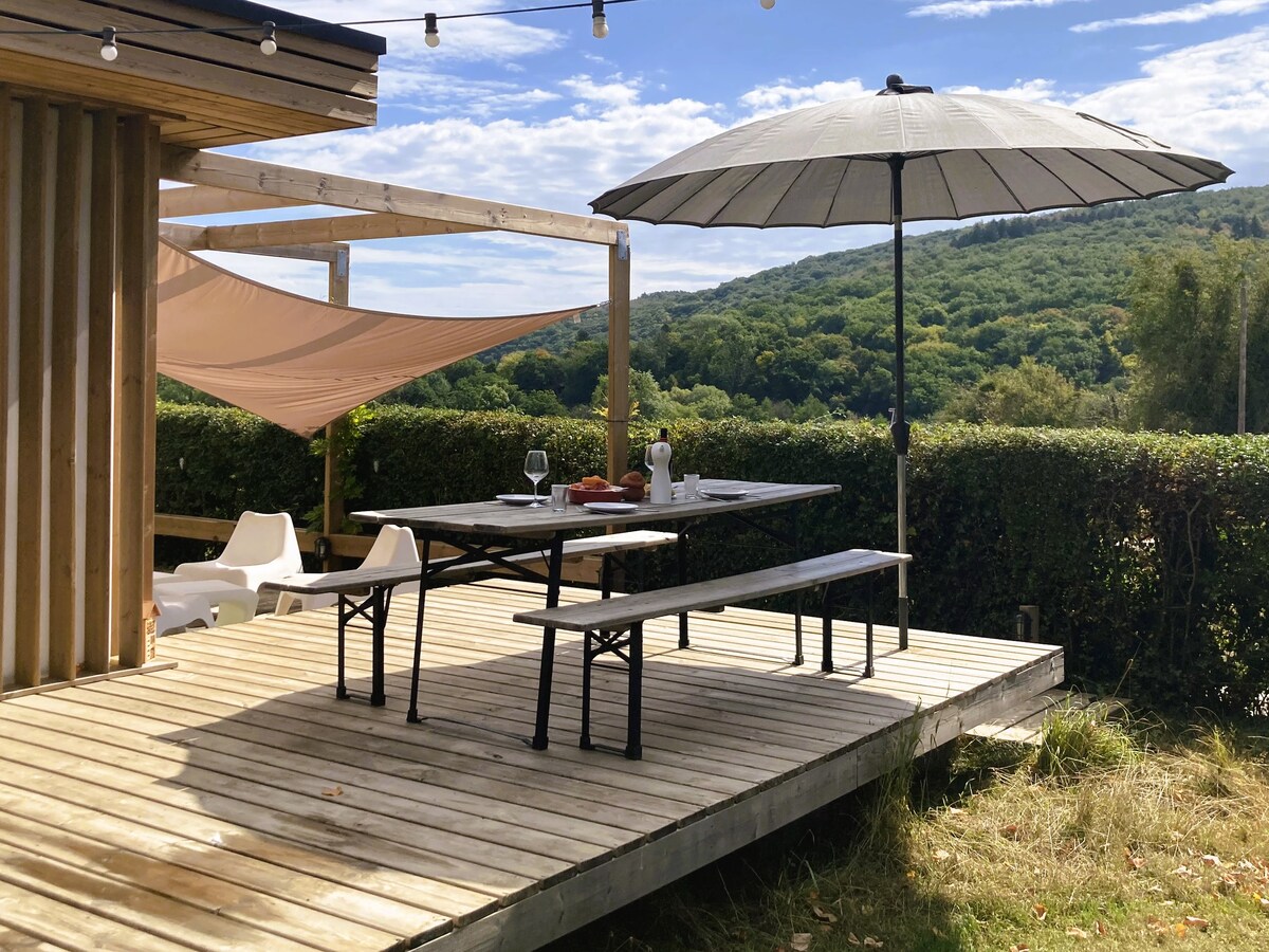 A wooden terrace offers a spacious dining area, featuring a large table beneath a wide umbrella. The setting showcases scenic views of lush greenery, with a relaxing atmosphere enhanced by the surrounding natural landscape.