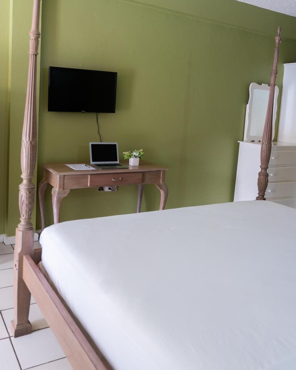 A spacious sleeping area features a four-poster bed with a neutral-toned mattress. A small desk with a laptop and a potted plant is positioned nearby. A flat-screen TV is mounted on the green wall, enhancing the room's functional layout.