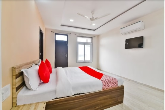 A bright sleeping area features a spacious bed with white linens and accentuated by red pillows. A wall-mounted television is visible across from the bed, and natural light enters through a large window. The walls are painted in a light neutral tone.