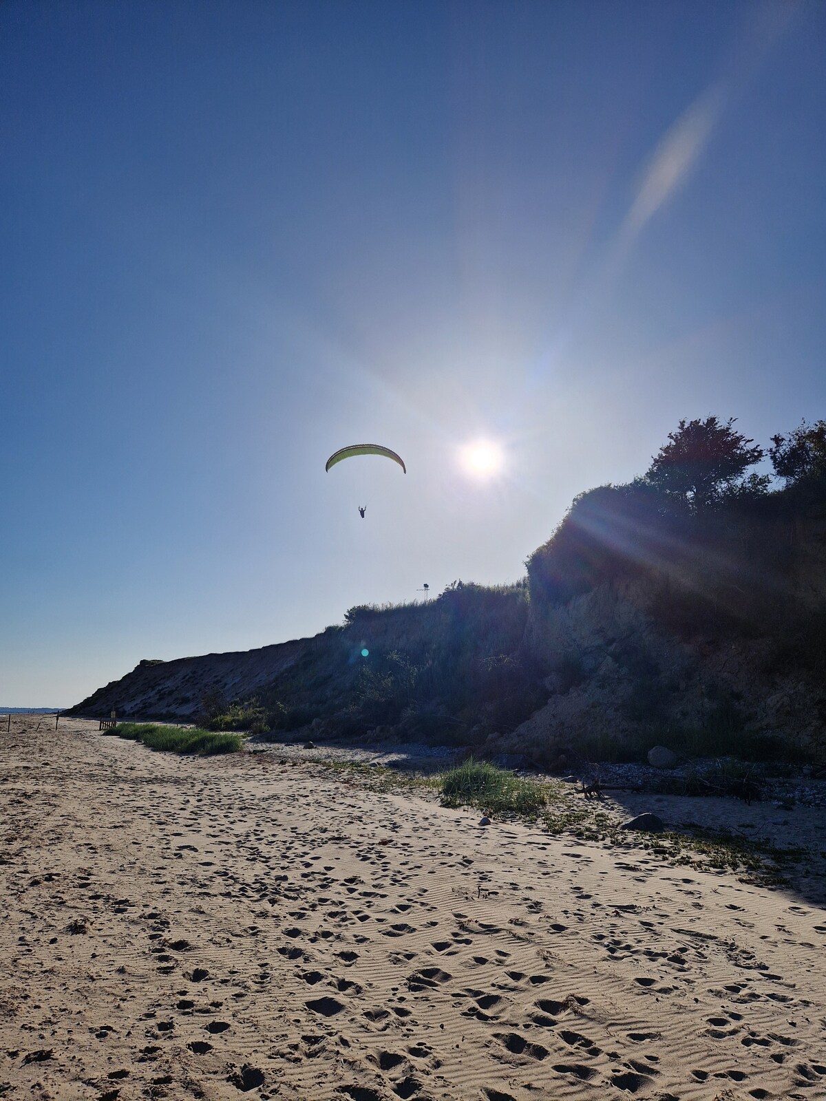 A clear sky is captured with a distant paraglider flying above a sandy beach. The sun is positioned low, casting a warm glow over the landscape. The shoreline features gentle undulations and patches of grass, with a natural setting in the background.