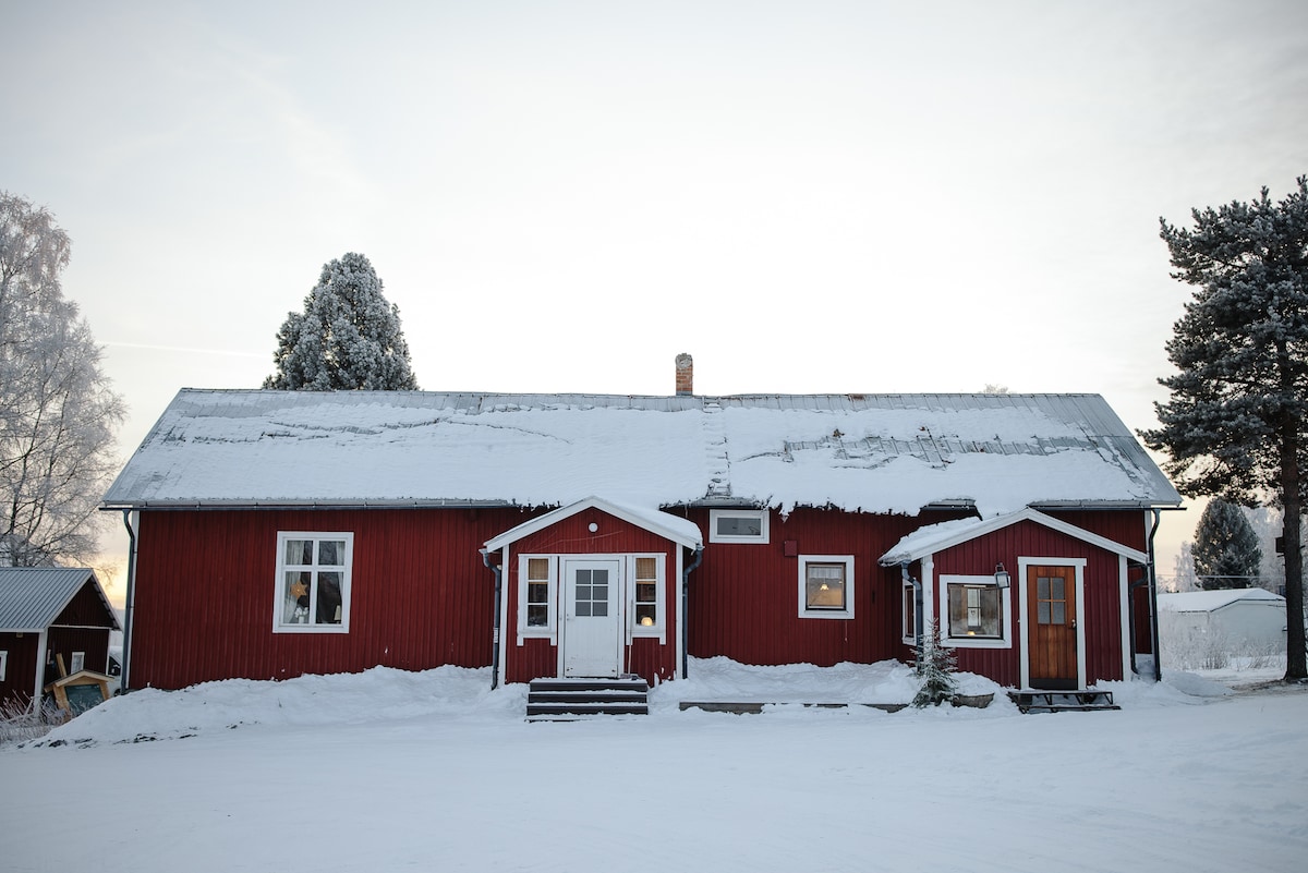 A charming red wooden villa is surrounded by snow, featuring multiple windows that allow for natural light. The entrance is marked by a set of steps leading to double doors, with additional structures visible nearby, adding to the cozy character of this winter setting.