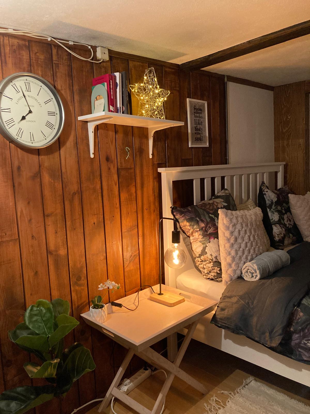A cozy sleeping area features a white bed adorned with decorative pillows and a soft blanket. A small table beside the bed holds a stylish lamp and a plant. A clock on the wooden wall adds functionality, while books are displayed on a nearby shelf.