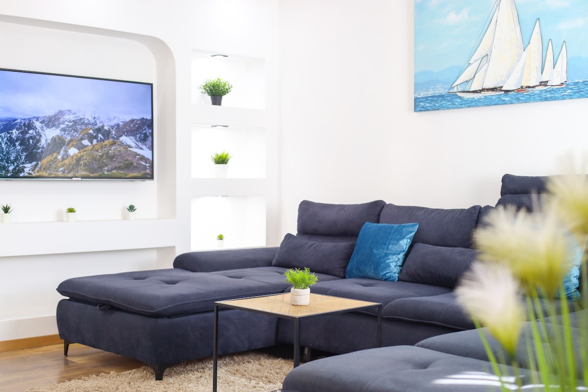 A modern living area is displayed with a dark sectional sofa featuring a light blue cushion. A flat-screen TV is mounted on the wall, complemented by decorative shelves with small plants. A coffee table sits at the center on a plush area rug, enhancing the room's comfort.