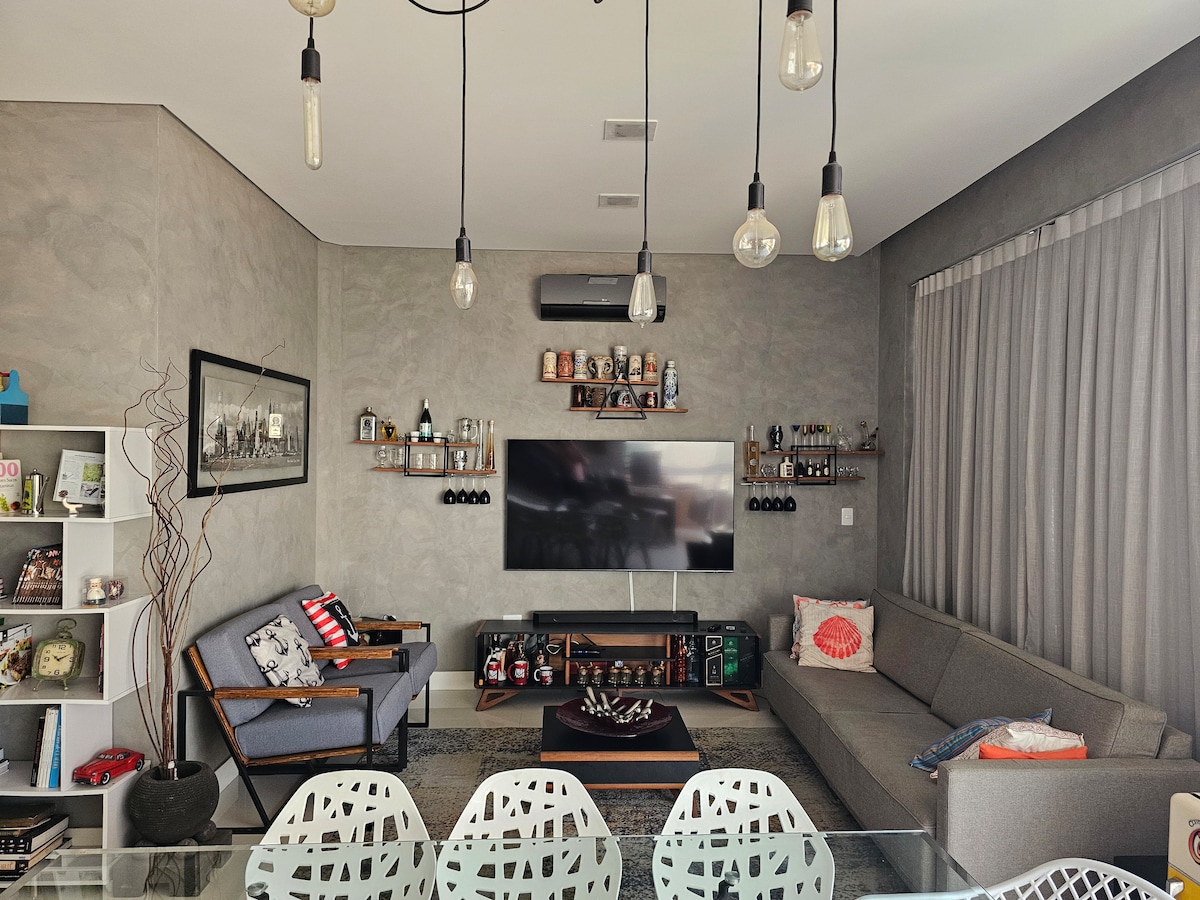 A modern living area features a gray sectional sofa and a minimalist coffee table. A wall-mounted television is positioned above a wood media unit, surrounded by shelves displaying decorative items. Large windows with sheer curtains allow natural light to fill the space, complemented by industrial-style pendant lights.