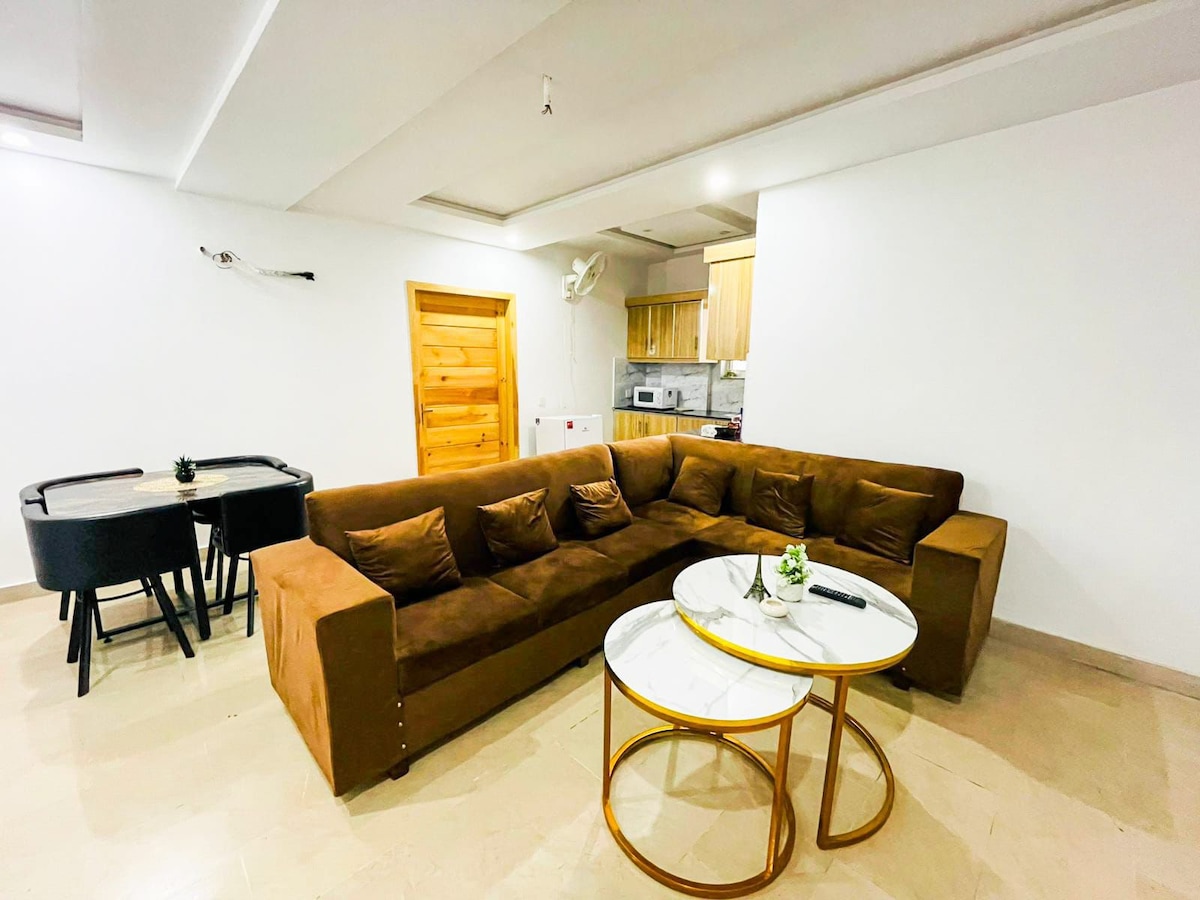 A spacious living area contains a large brown sectional sofa facing two round coffee tables. A dining area with a black table and four chairs is visible in the background, alongside a kitchen area featuring wooden cabinetry.