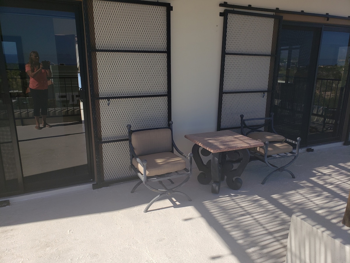 A private outdoor terrace is featured, showcasing a small wooden table surrounded by two comfortable chairs. Large sliding glass doors provide access to the interior while allowing natural light to brighten the space. Sunlight casts soft shadows across the light-colored flooring.