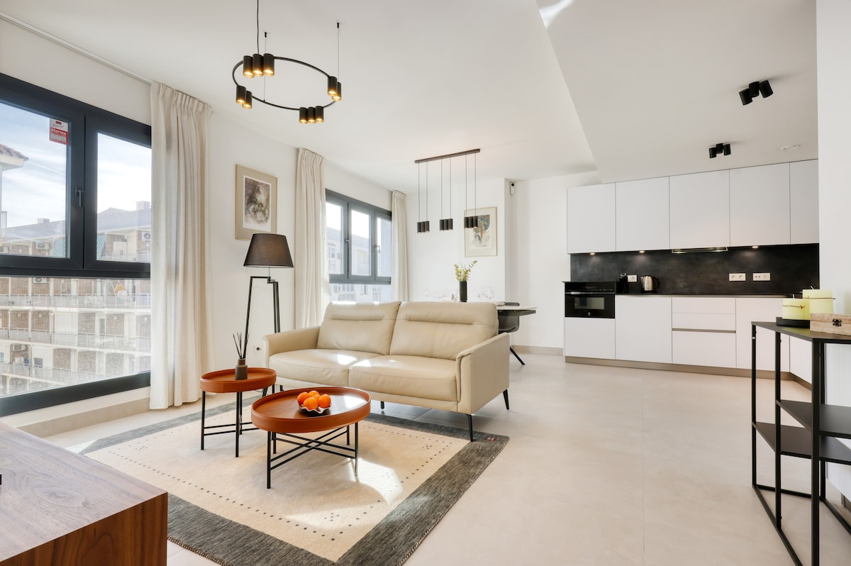 An open-plan living space is shown, featuring a light-colored sofa and modern coffee tables. A fully equipped kitchen is visible with sleek cabinetry. Large windows allow natural light to fill the room, enhancing the airy atmosphere and providing views of the surroundings.