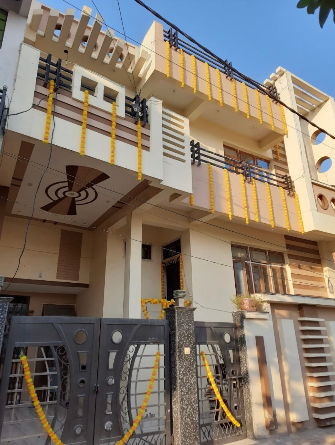 The exterior of a three-story building is showcased, featuring a modern façade with decorative elements. The entrance gate is adorned with traditional marigold garlands, while balconies are highlighted with contemporary railing designs and large windows allowing natural light.