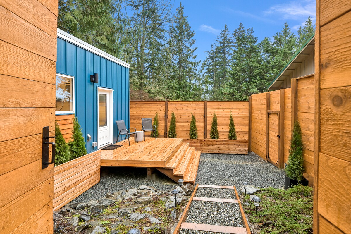A private outdoor area is framed by natural wooden fencing, featuring a cedar deck with seating for relaxation. Lush greenery surrounds the space, enhancing the tranquil forest atmosphere. A gravel pathway leads through the area, inviting exploration and enjoyment of the serene outdoors.