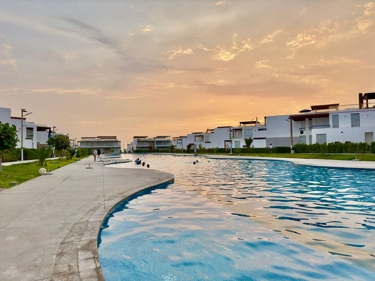 A serene swimming pool stretches alongside modern buildings under a gradient sky at sunset. The pool features a gentle curve, with a wide walkway bordered by lush greenery, inviting relaxation and leisure.