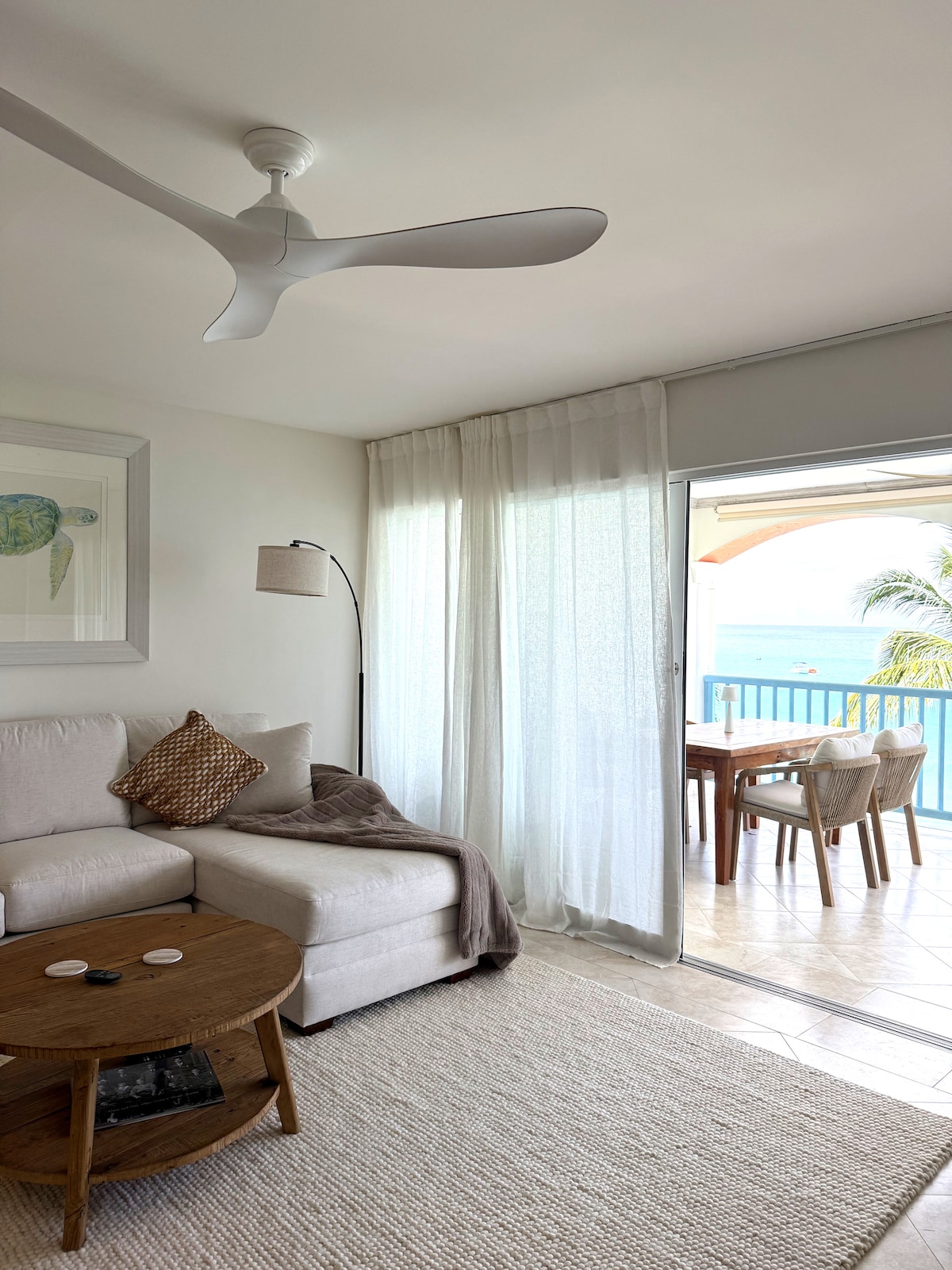 A light and airy living area features a white sofa adorned with a textured pillow, accompanied by a round wooden coffee table. Sheer curtains gently diffuse sunlight, while a door opens to a patio with a dining table overlooking the sea.