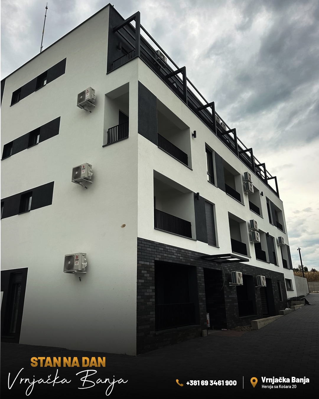The exterior of a modern three-story building is displayed, featuring a sleek white façade and black accents. Multiple air conditioning units are positioned on the walls, and dark balconies offer spaces for relaxation. A paved walkway leads to the entrance.