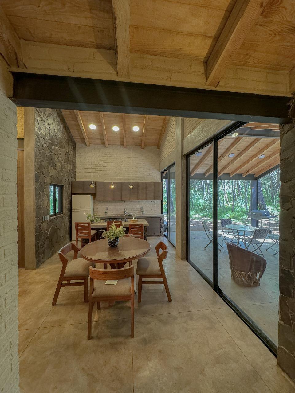 A dining area is featured, showcasing a round wooden table surrounded by chairs. Large glass doors allow natural light to fill the space, offering views of the forest outside. The ceiling displays wooden beams, enhancing the cabin's rustic charm.