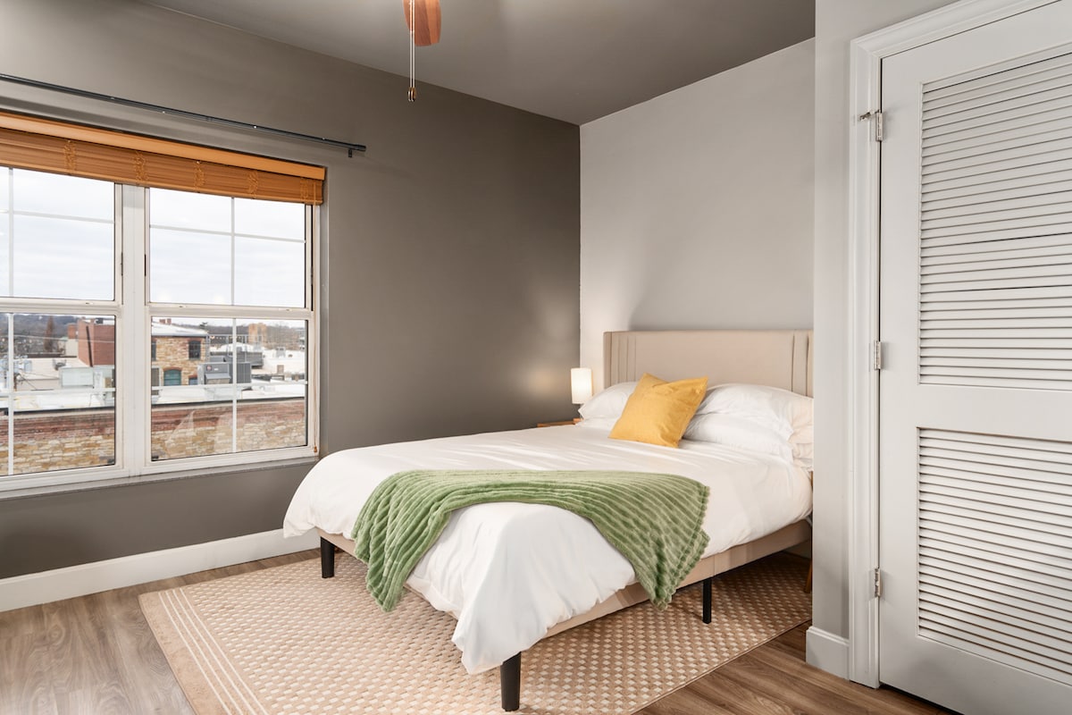 A well-appointed sleeping area features a queen bed topped with soft linens and a decorative yellow pillow. A green throw is casually draped over the bed. Natural light streams through two large windows, framed by light wooden blinds, enhancing the room's neutral decor.