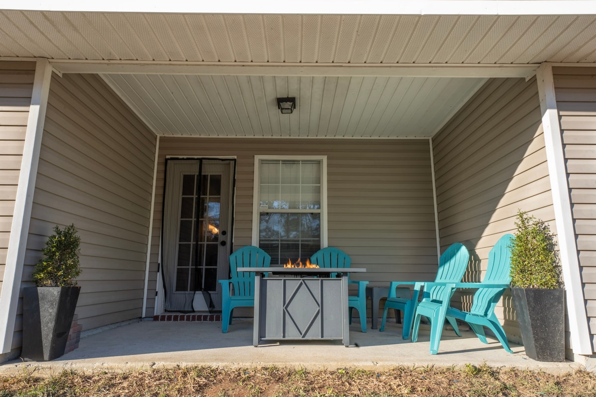 An outdoor seating area features a gas-powered fireplace at the center of a patio. Surrounding the fireplace, four turquoise chairs and a table create a cozy gathering space. Potted plants are positioned on either side, adding greenery to the area.