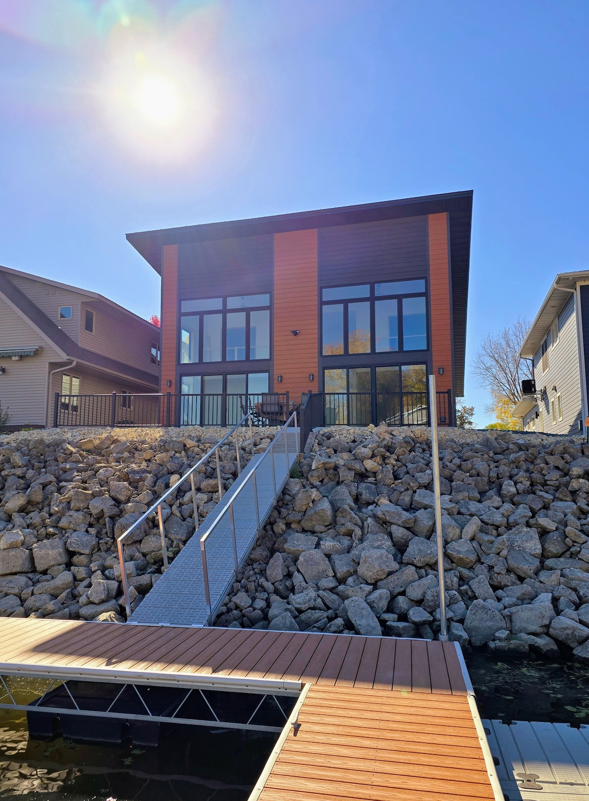 A modern two-story home features large glass windows and a contemporary wooden exterior. The structure is set against a rocky hillside, with a ramp leading down to a private dock on the water. Sunlight brightens the scene under a clear blue sky.