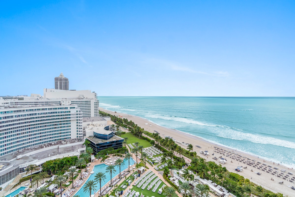 An expansive view of the oceanfront stretches across the horizon, showcasing gentle waves and a sandy beach. The scene includes an inviting pool area surrounded by palm trees and sun loungers, with the Fontainebleau Hotel's distinctive architecture seen in the background.