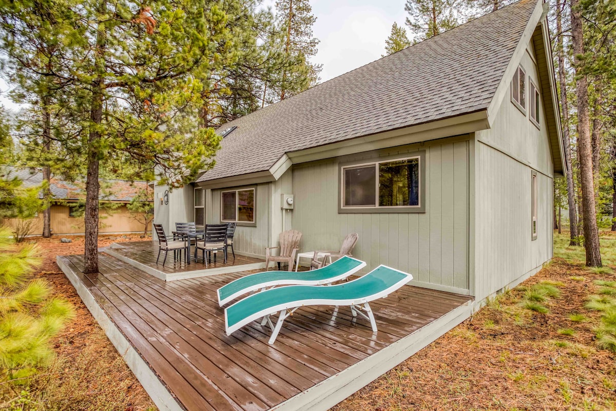 The exterior of a home is shown, featuring a spacious deck with lounge chairs. Surrounding trees provide natural shade, and a patio set is visible near the entrance. The design reflects a blend of indoor and outdoor living spaces, ideal for relaxation.