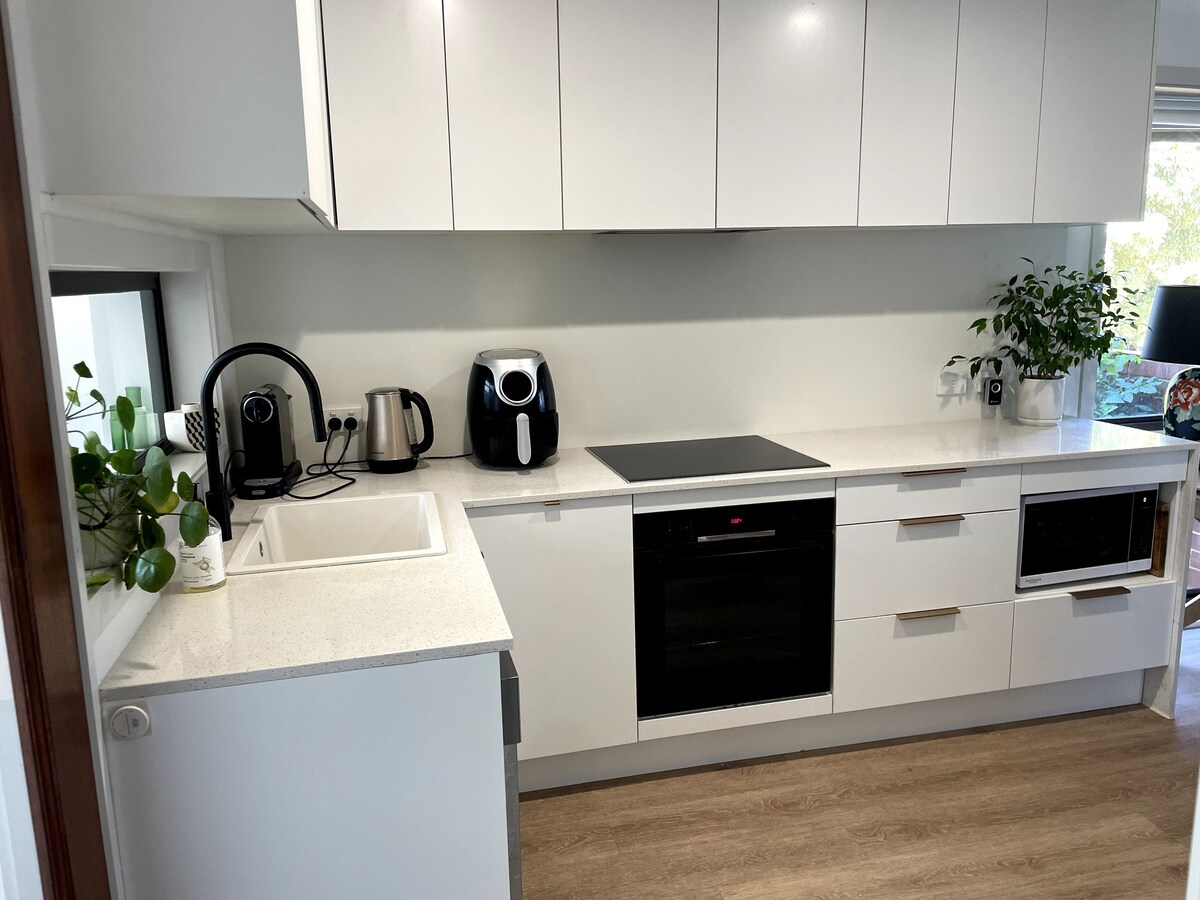 A modern kitchen is presented, featuring sleek white cabinetry and a light countertop. A variety of appliances are visible, including a coffee maker, air fryer, and built-in oven. Green plants add a touch of nature, contributing to the clean and organized appearance of the space.