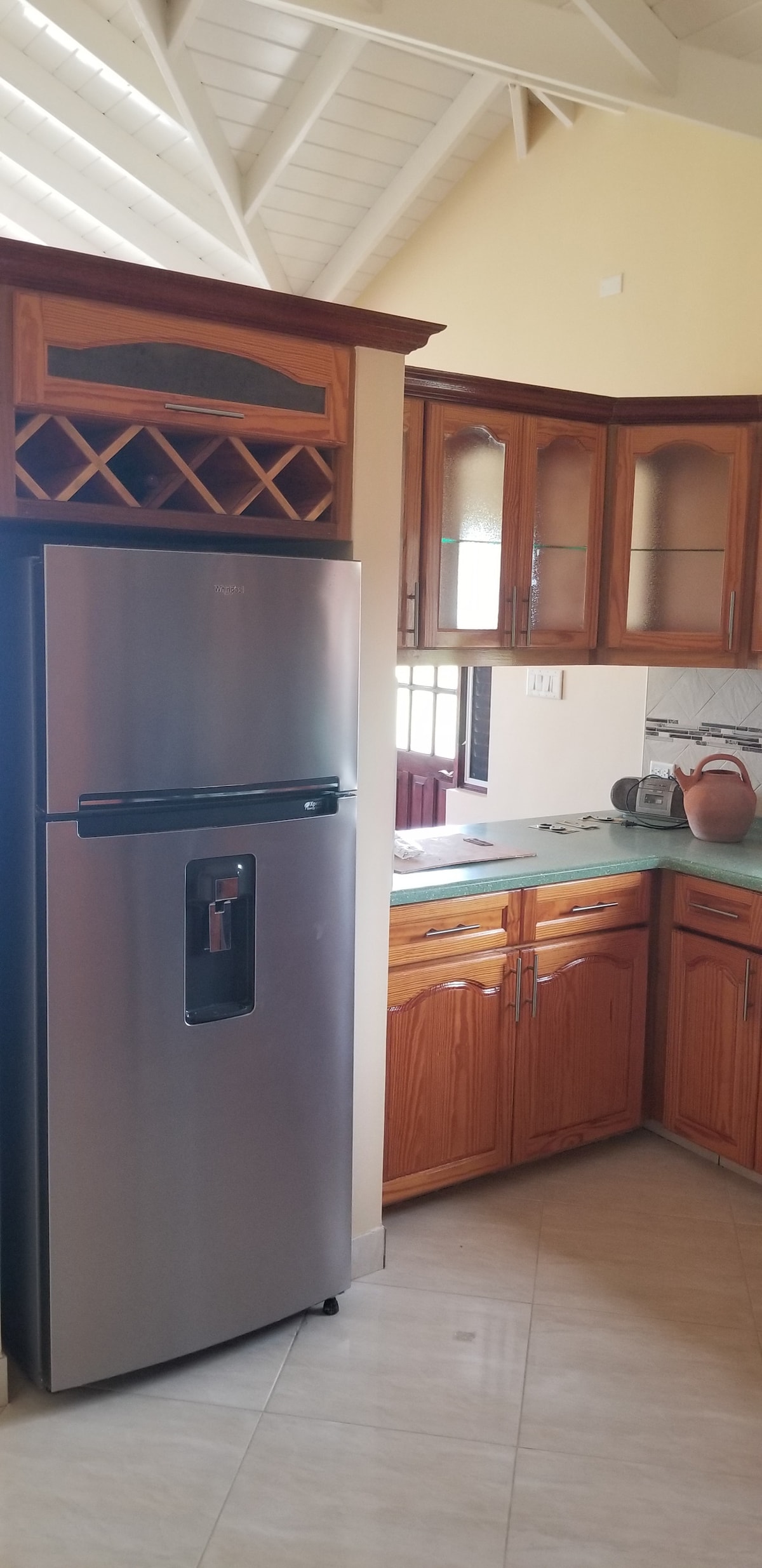A well-arranged kitchen features wooden cabinetry and a spacious countertop. A stainless steel refrigerator, equipped with a water dispenser, is positioned next to a wine rack. Natural light filters in through a window, creating a bright and functional cooking space.