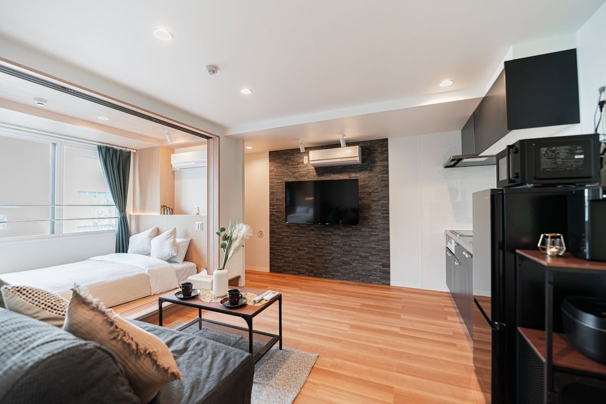 A modern living space features a comfortable sofa and a coffee table arranged in front of a wall-mounted television. Light wood flooring complements the open layout, which includes a spacious bed area separated by sliding doors. The kitchen area is equipped with essential appliances.