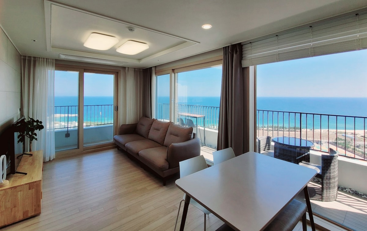 A bright living area features large windows offering panoramic ocean views. A spacious gray sofa is positioned next to a minimalist dining table. Natural light fills the room, creating a relaxed atmosphere, while the balcony highlights the coastal scenery beyond.
