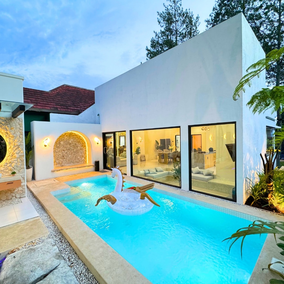 A serene outdoor area is highlighted by a rectangular pool, featuring a floating swan-shaped float. Large glass windows provide a view into the interior living space, which is filled with natural light. Lush greenery surrounds the pool, enhancing the tranquil atmosphere.