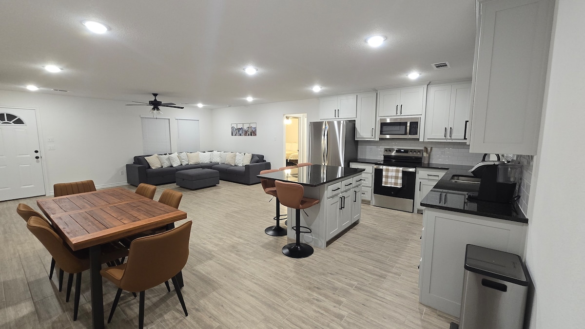 An open-concept living area features a spacious kitchen with modern appliances, including a refrigerator and oven. A dining table with six chairs is placed next to a large kitchen island with two bar stools. A comfortable sectional sofa is arranged nearby, creating a welcoming space.