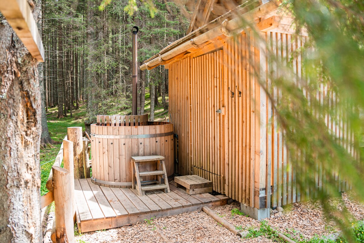A private wooden hot-tub is nestled beside a rustic wooden structure, surrounded by lush greenery. The hot-tub is crafted from timber, featuring a small wooden step leading up to it and is positioned on a wooden deck, integrating seamlessly with the natural surroundings.