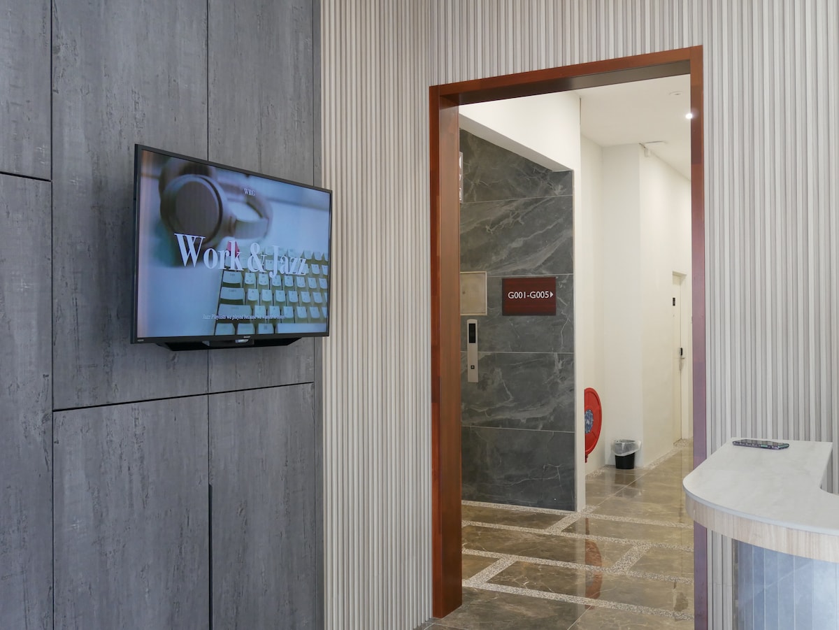 An entrance area is shown, featuring a wall-mounted television displaying the text 'Work & Play.' Smooth textured walls in neutral tones are complemented by a doorway leading to a corridor marked with signage. Reflective surfaces and polished flooring contribute to the modern design.