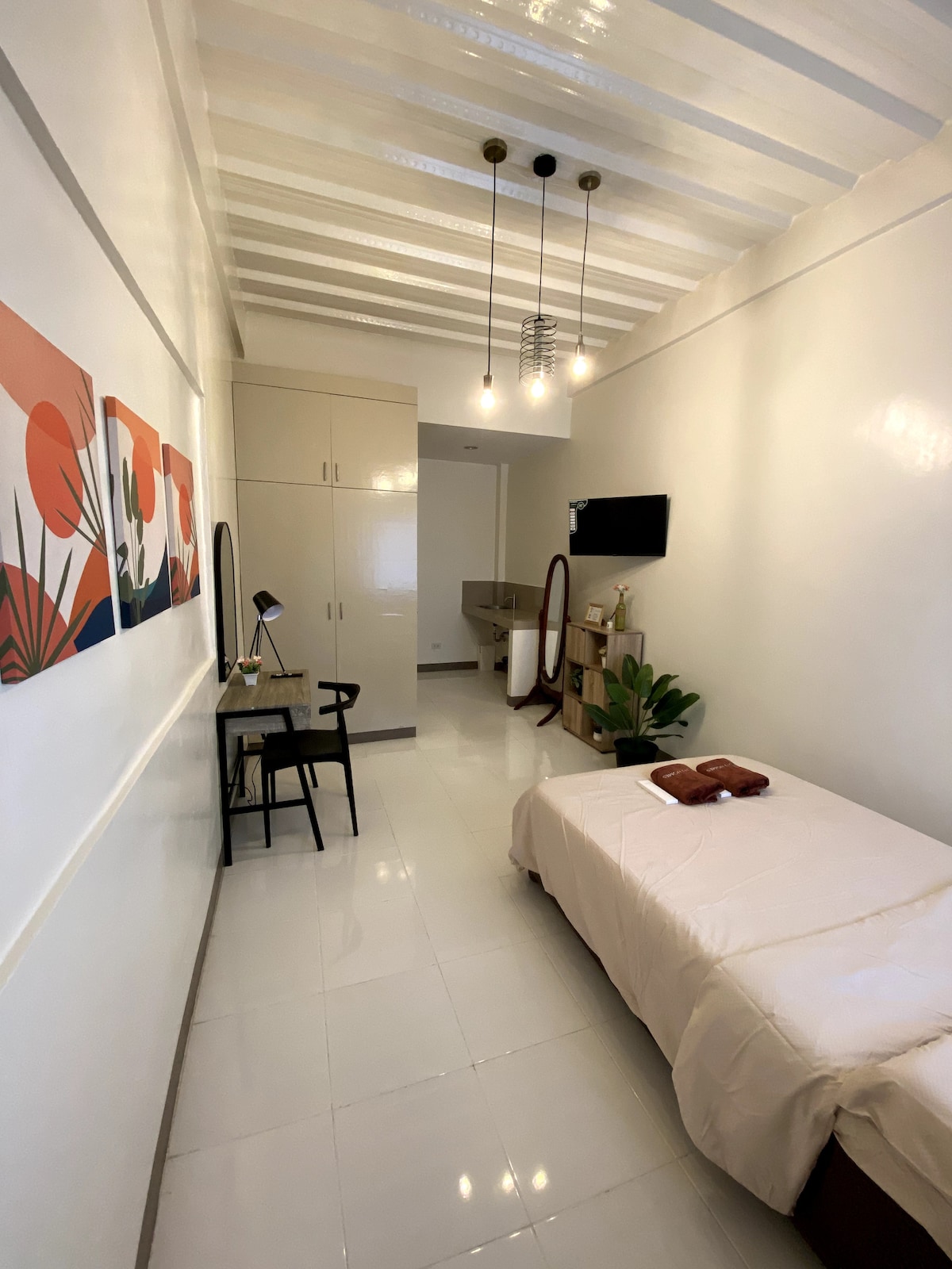 A bright room features a compact bed with a light-colored bedding set. An area with stylish artwork adds a pop of color to the white walls. A desk with a lamp and chair is positioned near a small kitchen area. Plants provide a touch of greenery.