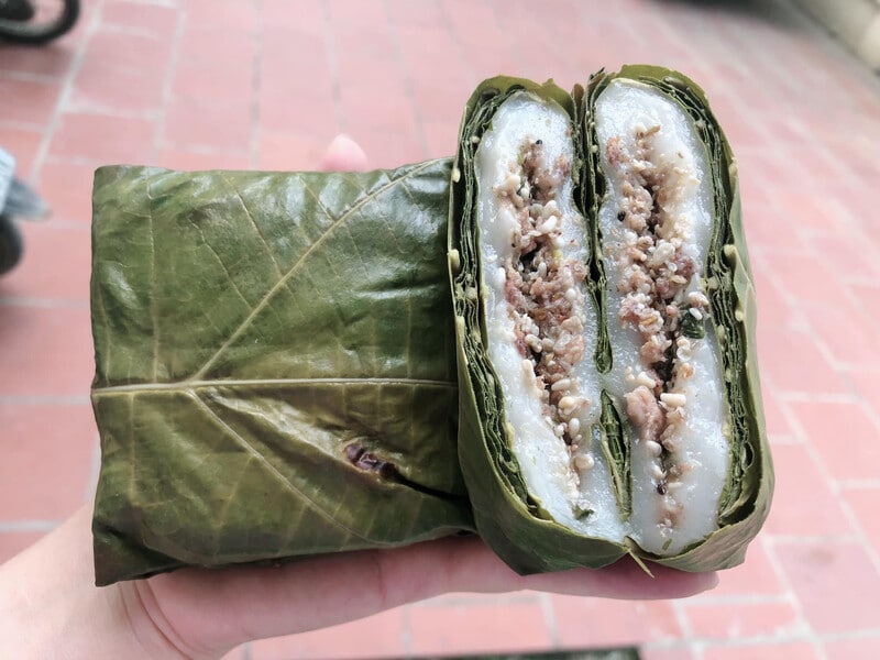 A wrapped food item is presented, showing layers of sticky rice and filling encased in large green leaves. The visible cross-section highlights a combination of meat and rice, neatly arranged, showcasing a traditional culinary preparation.