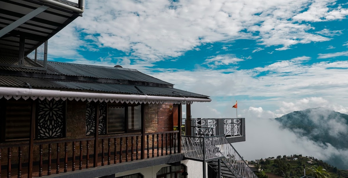 The wooden exterior of the cabin is highlighted against a backdrop of clear skies and rolling clouds. A balcony with intricate railing is visible, complemented by a staircase leading to the upper levels, offering access to stunning mountain views.