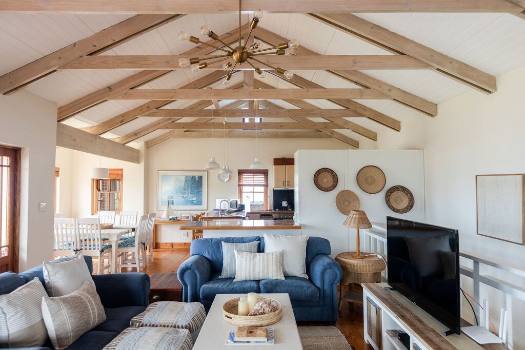 An inviting open-plan living area is highlighted by exposed wooden beams and large windows. Comfortable sofas provide seating, complemented by a coffee table adorned with decorative items. A Smart TV is positioned within the space, while a modern kitchen and dining area are visible in the background.
