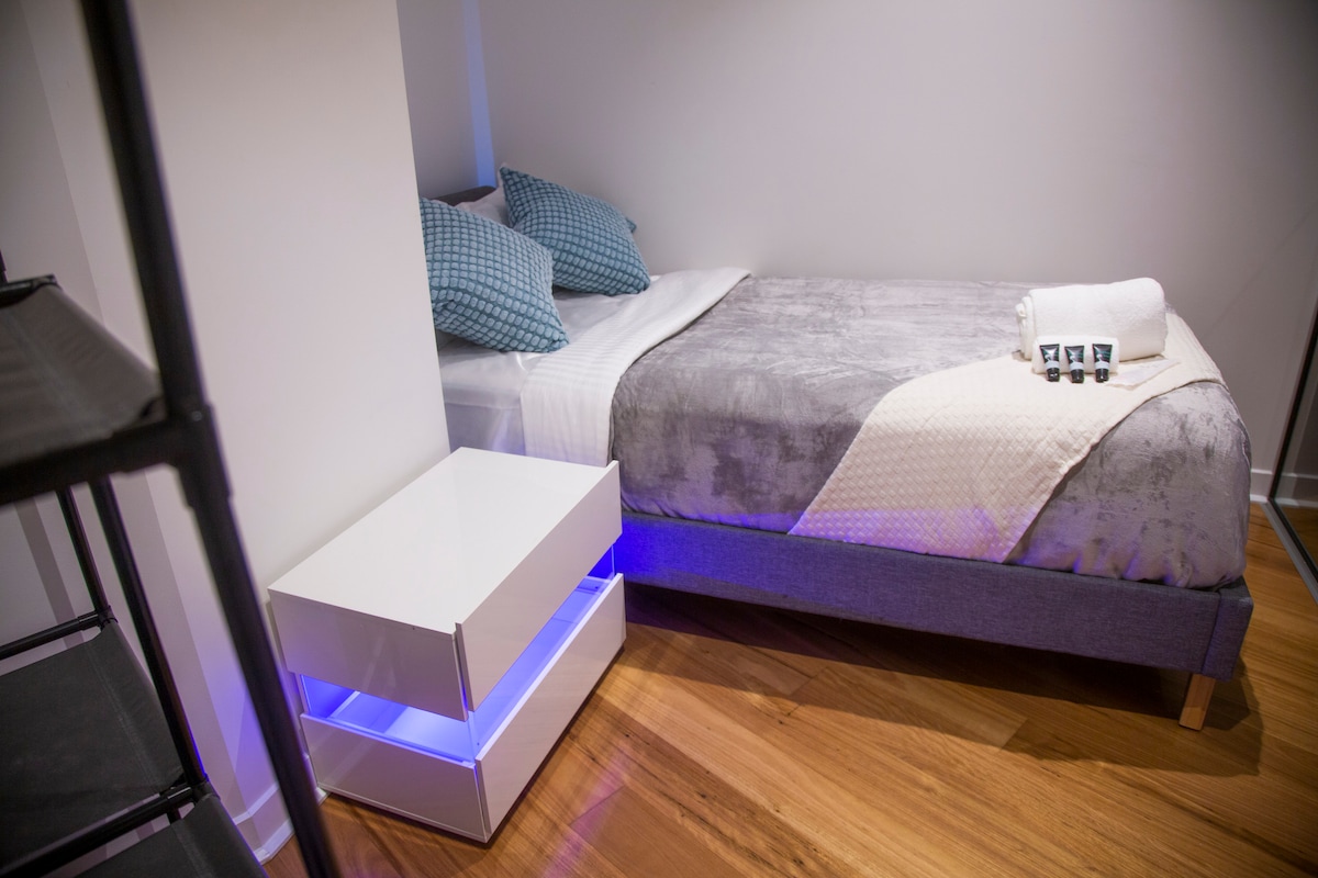 A private bedroom features a spacious bed with soft grey bedding. Two decorative blue pillows are placed at the head of the bed. A modern nightstand with under-lighting is positioned next to the bed, enhancing the contemporary design of the room.