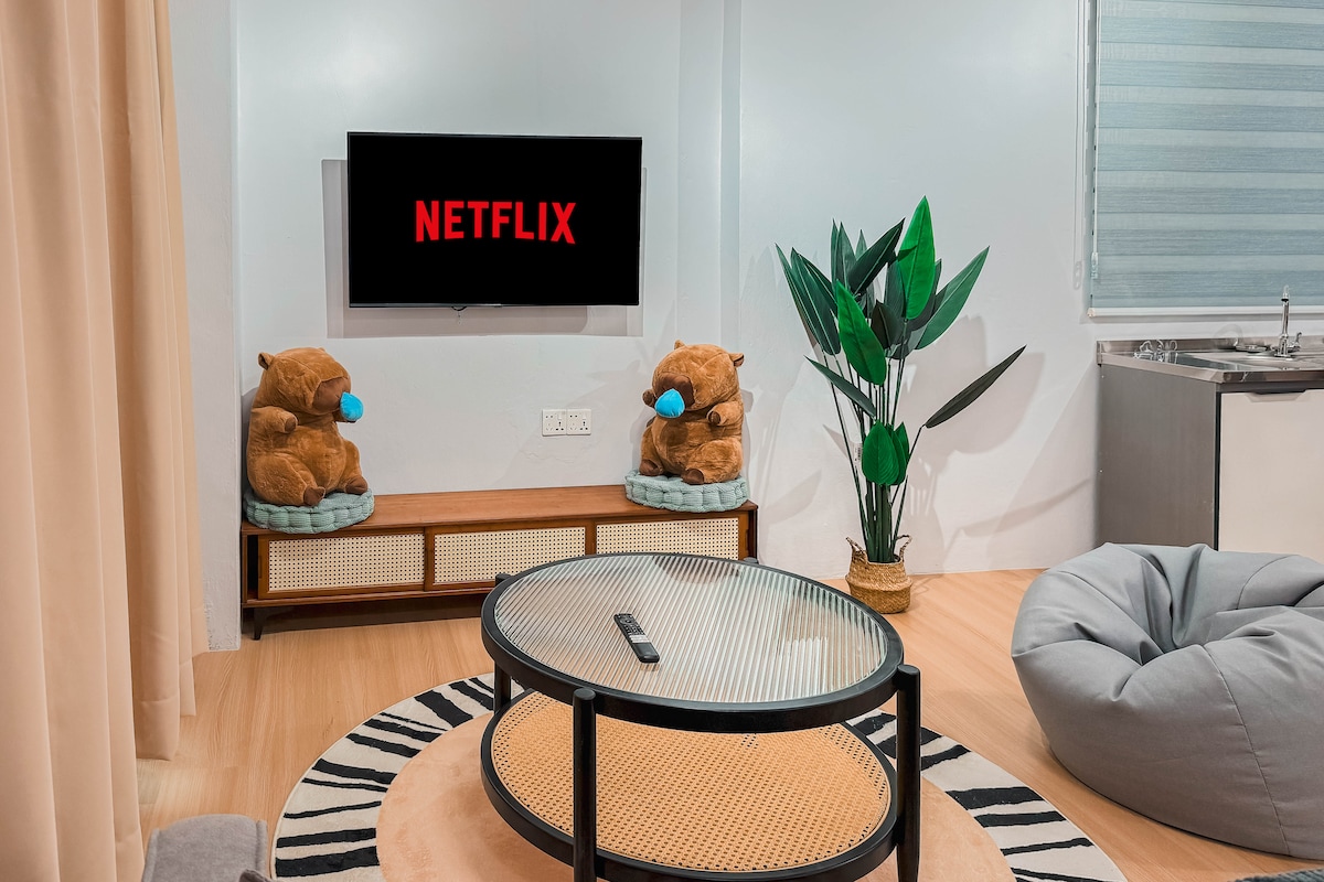 A cozy living area features a smart TV with a Netflix display centered above a wooden console. Two plush bear toys sit on either side. A round coffee table with a glass top is positioned on a patterned rug, alongside a soft bean bag chair and a green potted plant.