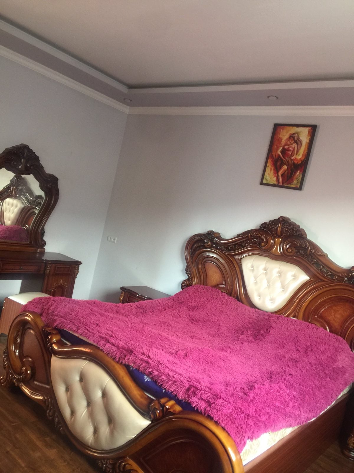 A spacious bedroom features an ornate wooden bed with a plush purple blanket draped over it. A matching wooden vanity and a dresser complement the decor, while a colorful wall painting adds a touch of character to the room. Natural light illuminates the space.