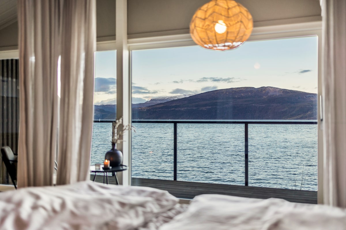 A serene view is presented from the large windows, showcasing the gentle waters of Ranfjorden and distant mountains. Soft curtains frame the scene, allowing natural light to fill the space. A decorative light fixture hangs above, adding a modern touch to the cozy interior.
