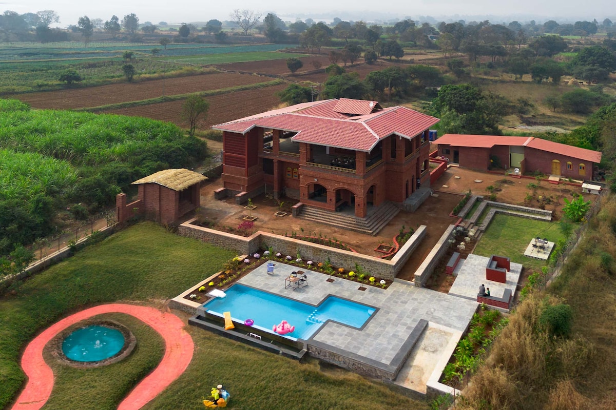 An aerial view of Bahava villa showcases its palatial facade surrounded by lush greenery and farmland. The outdoor pool and playful amenities can be seen in the backyard, with pathways leading through the landscaped garden. The traditional architecture is highlighted against a serene sky.