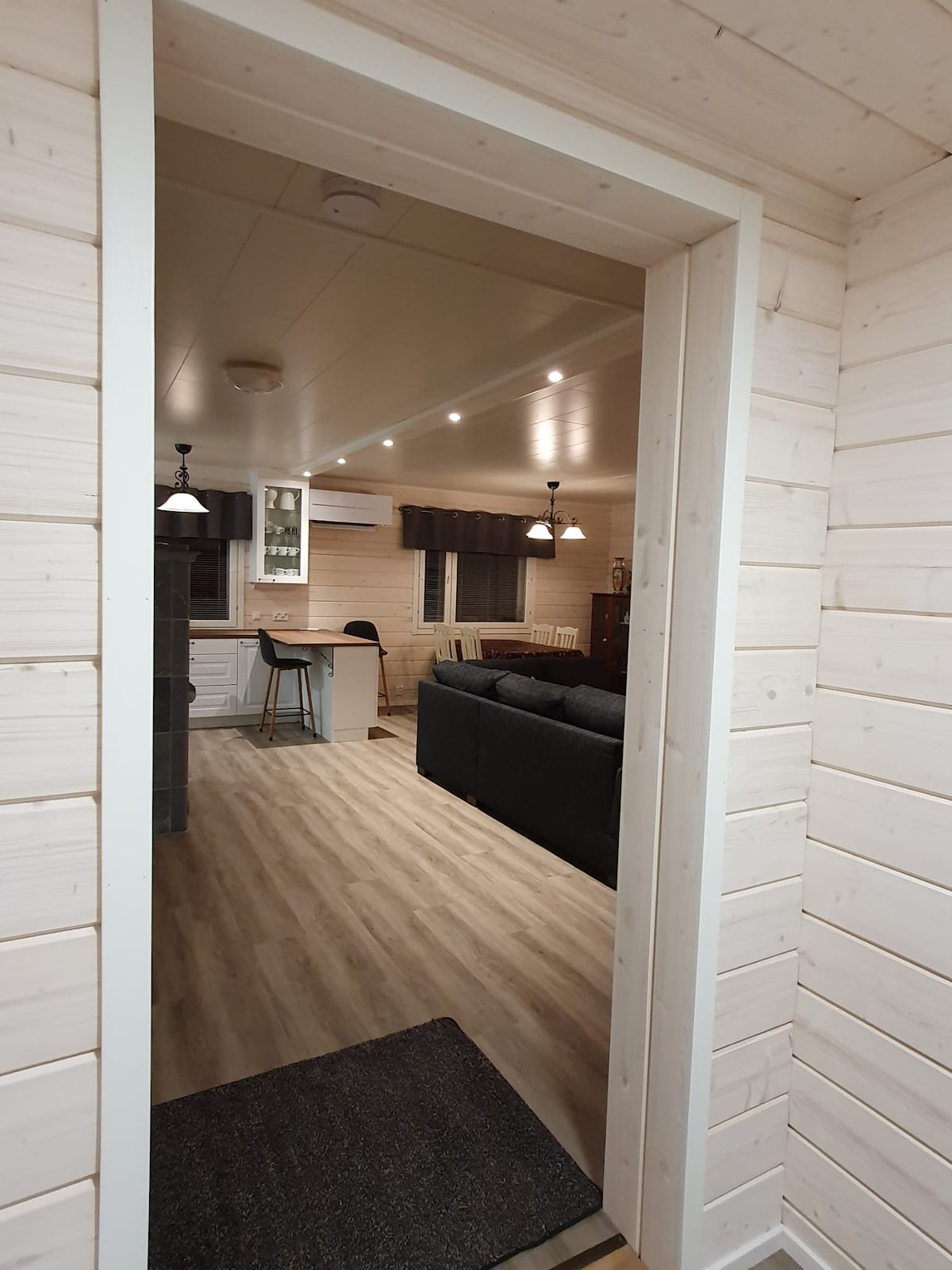 A view from a hallway showcases a modern, open living area featuring a dark sofa, a dining table, and a kitchen space. Recessed lighting illuminates the room, while light wood paneling creates a cohesive, inviting look. A doorway leads to the outside.