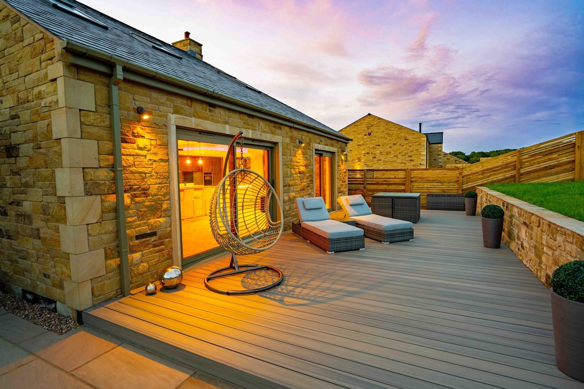 A spacious wooden deck is showcased, featuring a stylish hanging chair and two lounge chairs. Soft light illuminates the area, highlighting the warm tones of the stone exterior. The backdrop presents a serene landscape under a colorful sunset sky.