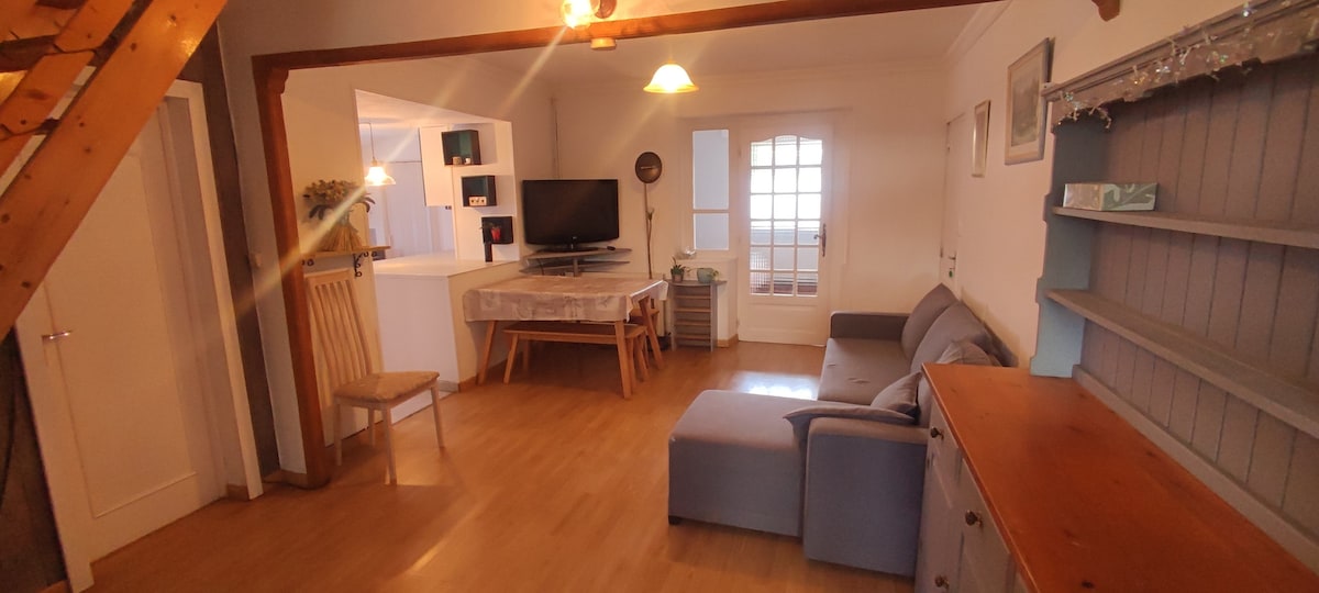 A spacious living area features a comfortable gray sofa and a wooden dining table. A television is mounted on the wall, and natural light enters through a set of glass doors. Decorative shelves and a wooden sideboard add functional elements to the room.