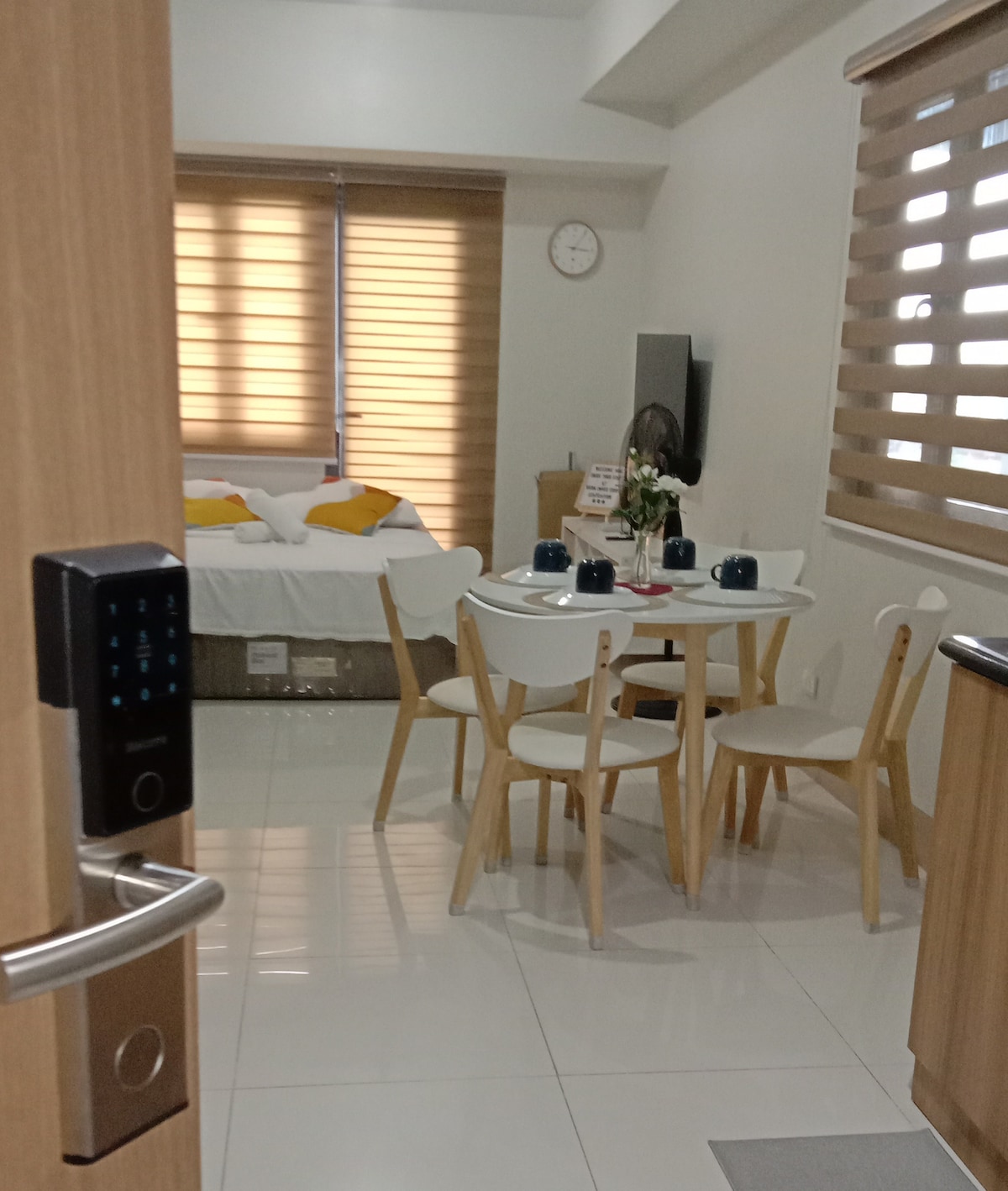 A bright interior space features a dining table with four white chairs. A spacious bed with yellow accent pillows is visible in the background. Window treatments provide light control, and a clock hangs on the wall, complementing a simple yet functional living area.