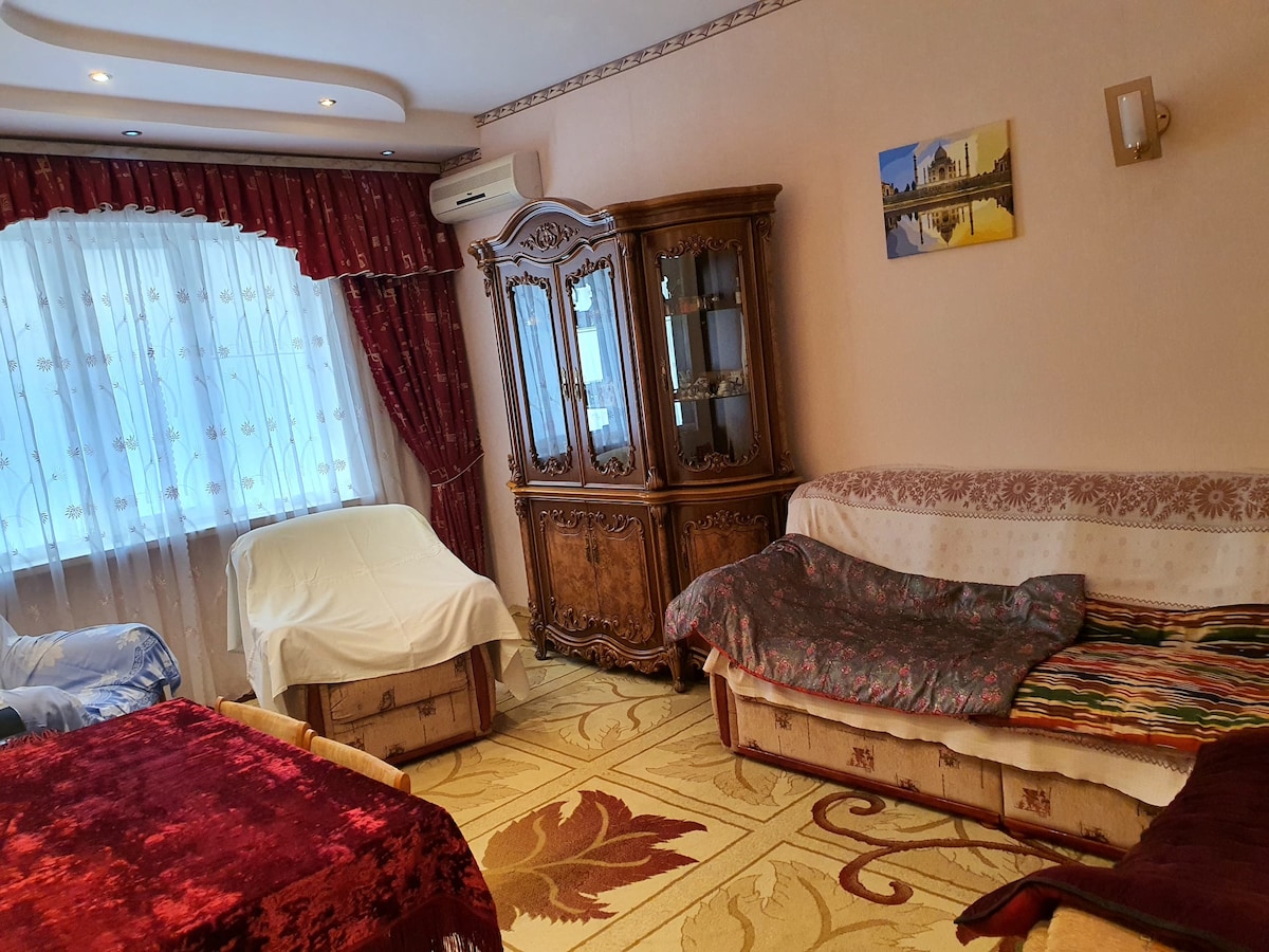 A warm living area is presented with two individual beds covered by patterned blankets. An ornate wooden cabinet stands prominently, with a framed artwork displayed above. Light filters through a window dressed in sheer curtains, contrasting with rich burgundy drapes.